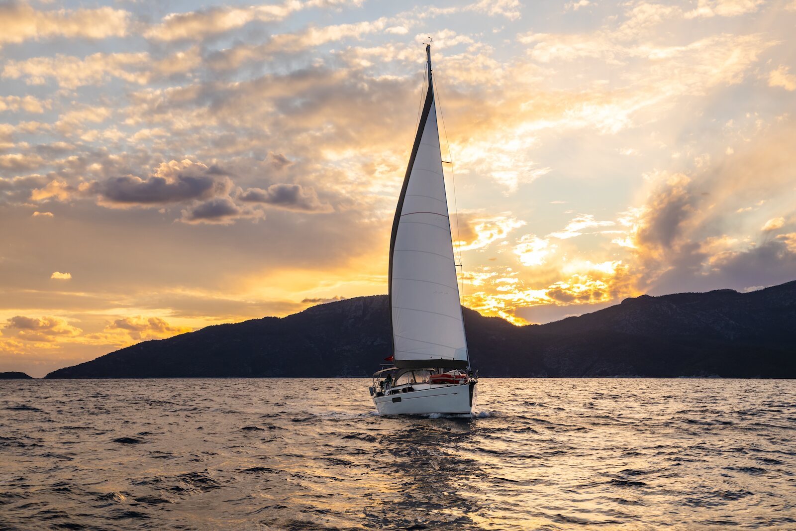 A white sailboat with full sails glides across open water at sunset, silhouetted mountains and a golden cloudy sky in the background